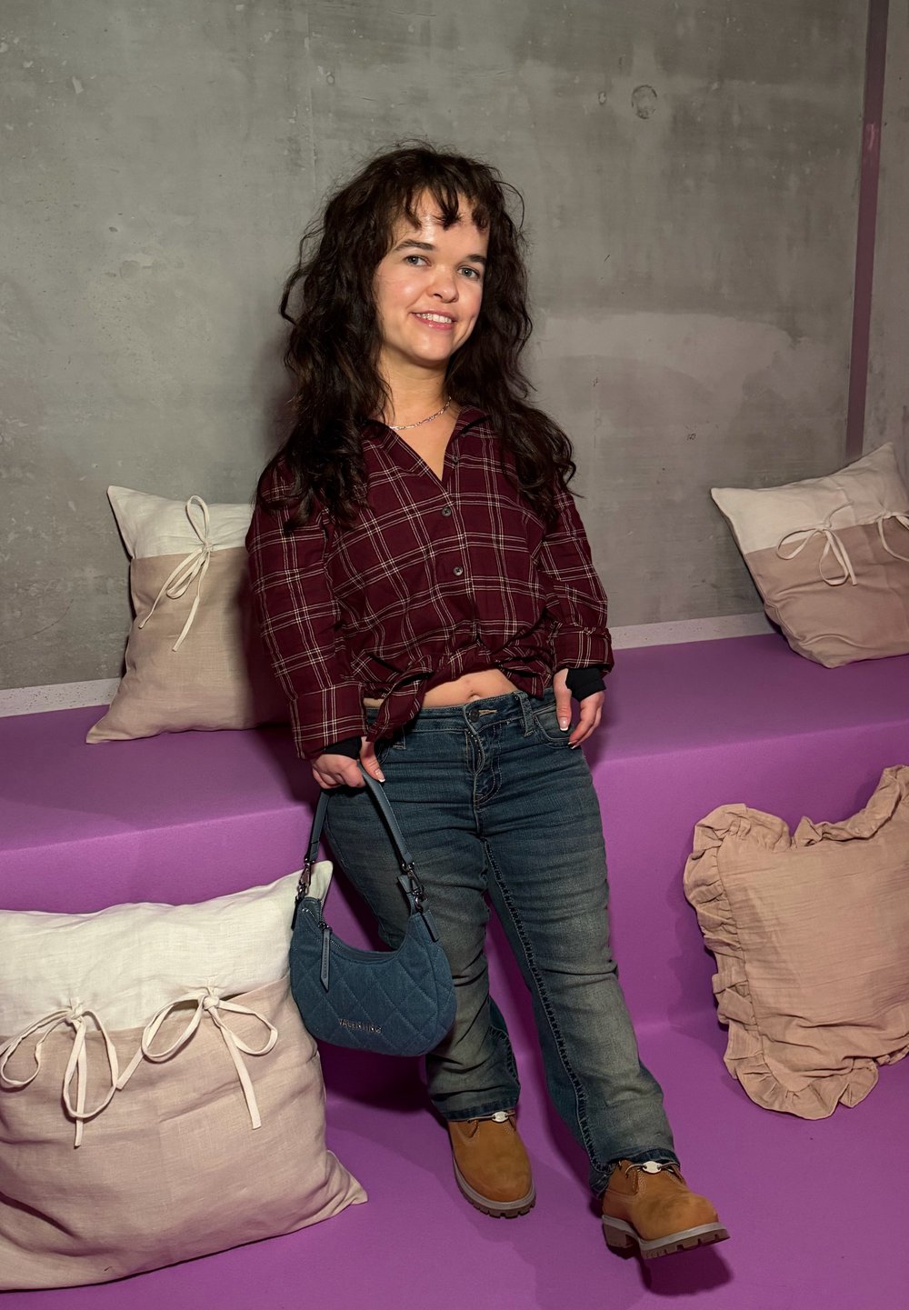 Femme souriante aux longs cheveux bouclés portant une chemise à carreaux rouge, un jean et des bottes marron, tenant un petit sac à main bleu dans une pièce avec des sièges violets.