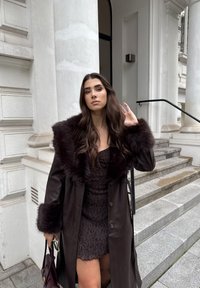 Brun skinnjakke med pelskrage, lagdelt over en mørk mønstret kjole. Teksturert stoff med en svak glans, komplementert av en håndveske.