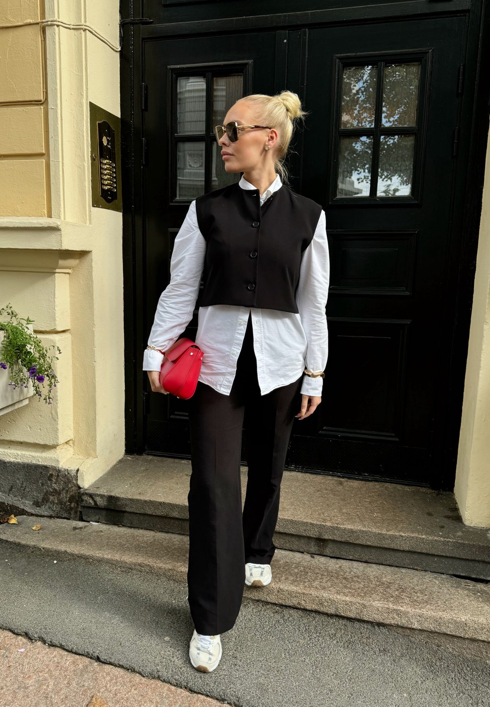 Μαύρο κοντό γιλέκο πάνω από άσπρο πουκάμισο με κουμπιά, μαύρο παντελόνι φαρδύ, και άσπρα αθλητικά παπούτσια. Κρατάει μία μικρή κόκκινη τσάντα. Φόντο με μαύρη πόρτα.