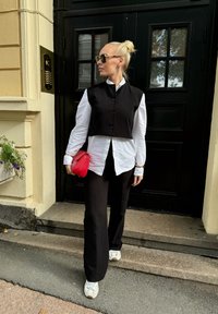 Μαύρο κοντό γιλέκο πάνω από άσπρο πουκάμισο με κουμπιά, μαύρο παντελόνι φαρδύ, και άσπρα αθλητικά παπούτσια. Κρατάει μία μικρή κόκκινη τσάντα. Φόντο με μαύρη πόρτα.