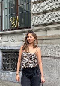 Leopardprint cami met dunne bandjes, gecombineerd met hoge zwarte jeans. De achtergrond bevat decoratieve steenarchitectuur en een bord.