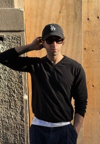 Suéter negro de manga larga, gorra gris con el logo "LA" y gafas de sol negras. De pie contra un fondo de madera con una pared texturizada.