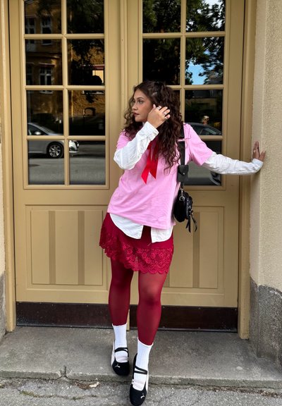 El modelo lleva una camiseta rosa, una camisa blanca con cuello debajo, una falda roja con encaje, medias rojas y zapatos negros con calcetas blancas, de pie junto a una puerta.