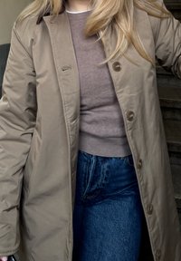 Lysebrun, knælængde frakke med knaplukning og blød tekstur, båret over en tætsiddende grå sweater og mørkeblå jeans.