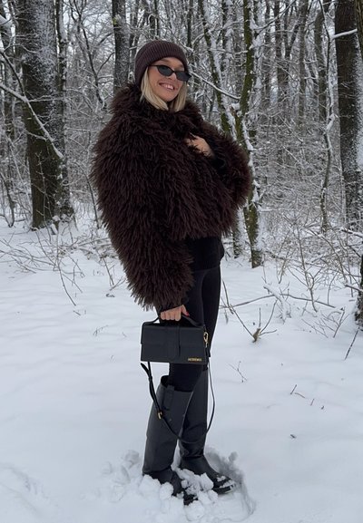 Chaqueta de piel sintética marrón, suéter negro, leggings negros, botas negras brillantes hasta la rodilla y bolso estructurado negro con detalles dorados. Fondo nevado.