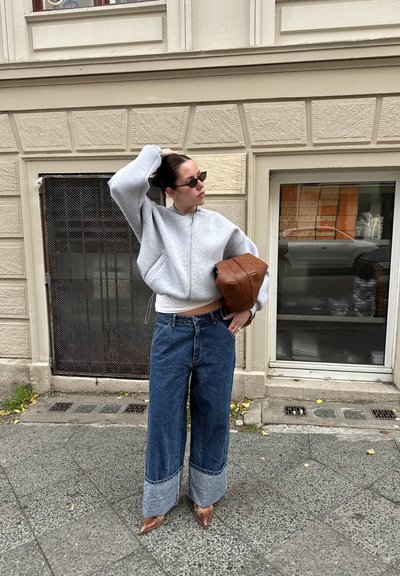 Sudadera gris con cremallera, vaqueros azul de pierna ancha con puños desgastados y bolso de cuero marrón. Los accesorios incluyen gafas de sol oscuras y un reloj.