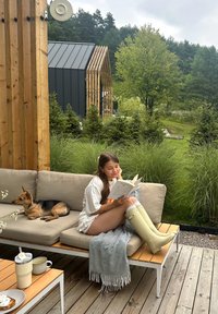 En person som sitter i hvite gummistøvler leser en bok på en beige sofa med et grått teppe. En liten hund slapper av ved siden av dem. Grønne trær i bakgrunnen.