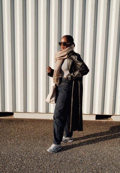 Abrigo largo de cuero negro, camiseta ajustada gris, pantalones de mezclilla oscuros, zapatillas gris, bufanda beige y gafas de sol. De pie contra un fondo blanco texturizado.