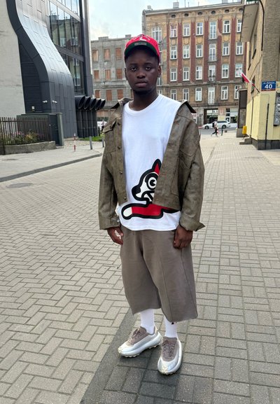 Chaqueta marrón con un patrón, camiseta blanca con un gráfico de un gato negro y rojo, pantalones cortos beige holgados, calcetines blancos y zapatillas gruesas grises.