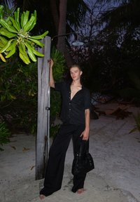 Pessoa vestindo camisa preta de botões e calças de perna larga, segurando um poste de madeira com um chuveiro, cercada por folhagem verde em uma área arenosa.