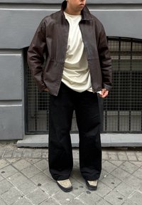 Barna bőrkabát cipzárral, krém színű póló és fekete, bő szárú nadrág. A lábbeli szürke felsőrésszel és fekete talpakkal rendelkezik.