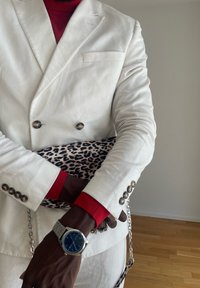 Osoba v bílém blejzru a červené košili, která drží kabelku s leopardím vzorem a stříbrným řetízkem, má na levém zápěstí stříbrné hodinky s modrým ciferníkem.