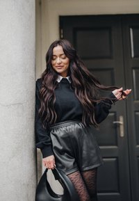 Femme aux cheveux longs et ondulés portant un pull noir, un short en cuir, des collants à motifs, tenant un sac à main noir, debout près d'une colonne et d'une porte sombre.