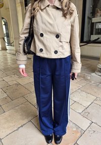 Casaco cropped de duas fileiras de botões em bege, calças de pajamas em cetim azul-marinho com riscas laterais brancas, mala preta e sapatos pretos com detalhes texturizados.