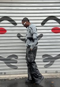 Suéter gris difuso con rayas negras; pantalones cargo de cuero negro holgados; gafas de sol blancas; de pie contra una persiana metálica decorada con grafitis.