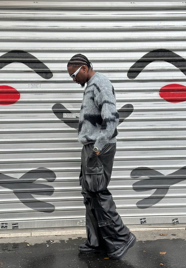 Harmaa pörröinen neule mustilla raidoilla; löysät mustat nahkaiset cargo-housut; valkoiset aurinkolasit; seisoo graffitilla koristetun metallisäleikön edessä.