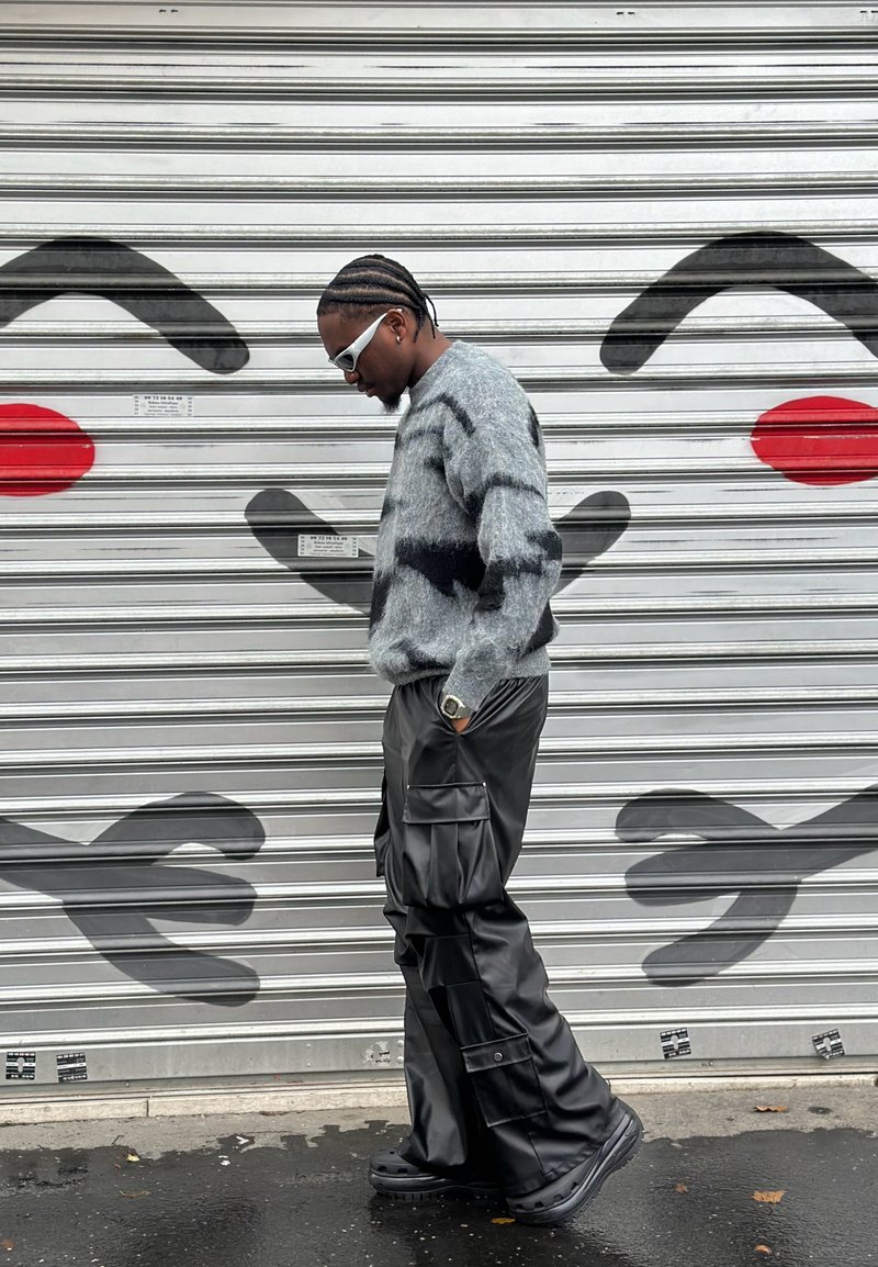 Grijze pluizige trui met zwarte strepen; losse zwarte leren cargobroek; witte zonnebril; staand tegen een met graffiti bedekte metalen rolluik.