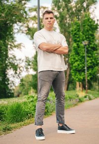 Ανοιχτόχρωμο μπλουζάκι, γκρι στενά τζιν και μαύρα αθλητικά παπούτσια με λευκές σόλες. Ο άνδρας στέκεται με τα χέρια σταυρωμένα σε ένα πάρκο.