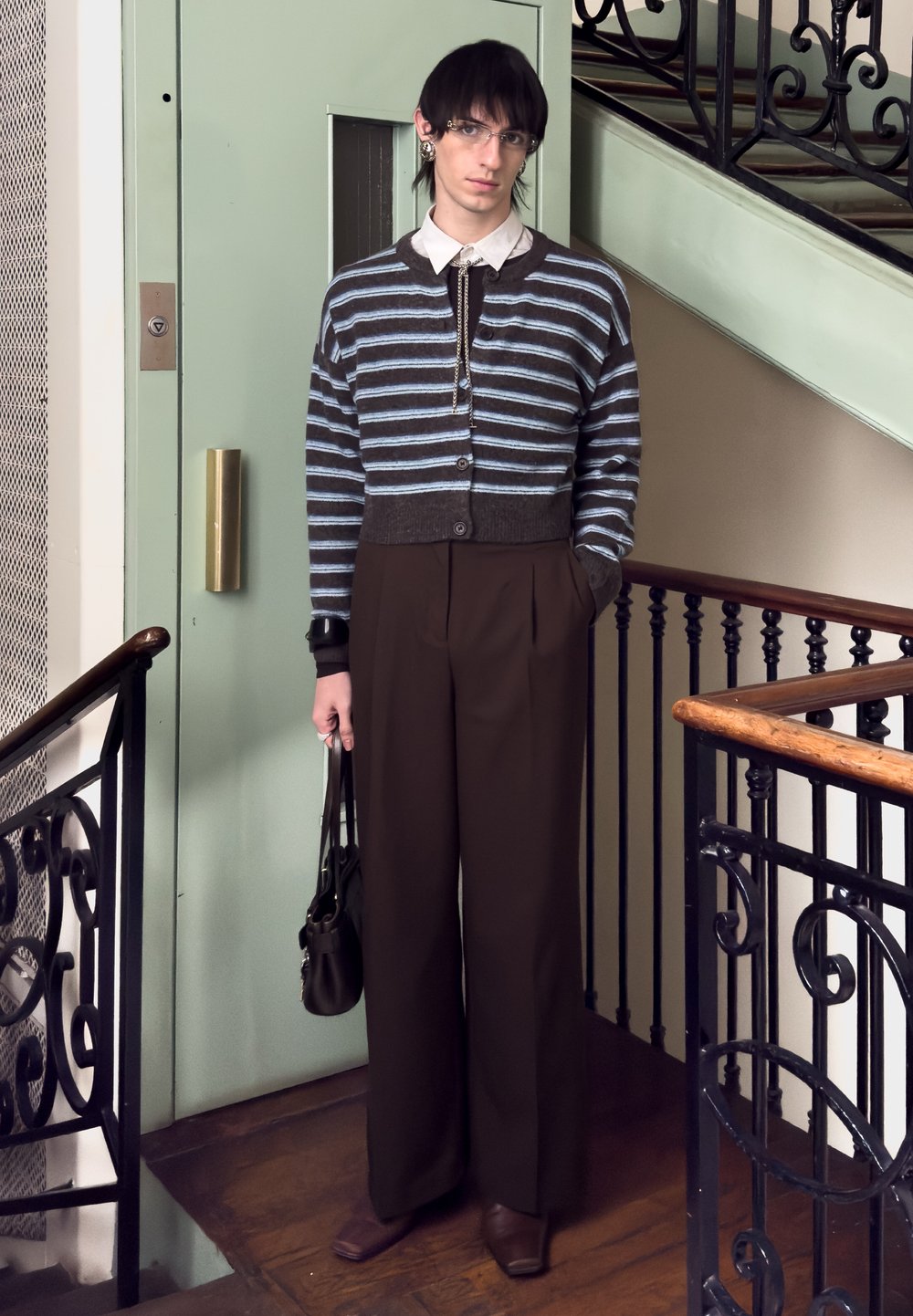 Jeune personne aux cheveux noirs courts et portant des lunettes, debout à l'intérieur près d'une rampe d'escalier, vêtue d'un cardigan rayé, d'un pantalon large et tenant un sac à main.