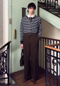 Jeune personne aux cheveux noirs courts et portant des lunettes, debout à l'intérieur près d'une rampe d'escalier, vêtue d'un cardigan rayé, d'un pantalon large et tenant un sac à main.