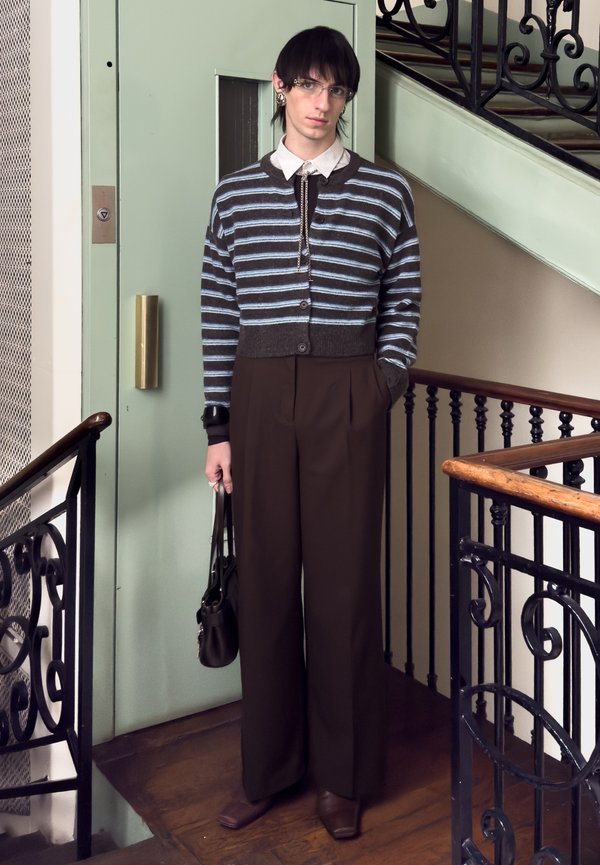 Ung person med kort svart hår og briller står innendørs ved et rekkverk, iført en stripete cardigan, vide bukser og holder en håndveske.