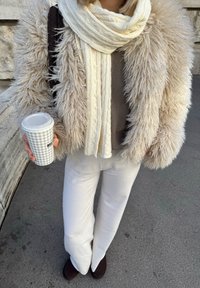 Pumpuli beige turkki, kerma värinen neulehuivi, harmaa paita, valkoiset housut, tummanruskeat kengät, kädessä kuvioitu kahvimuki.