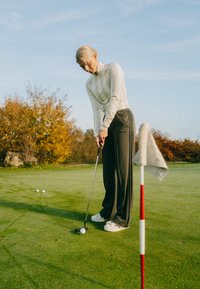 Golfer in einem hellen Langarmshirt und dunkler, weiter Hose, der einen weißen Golfball auf grünem Gras nahe einer rot-weißen Fahnenstange puttet.