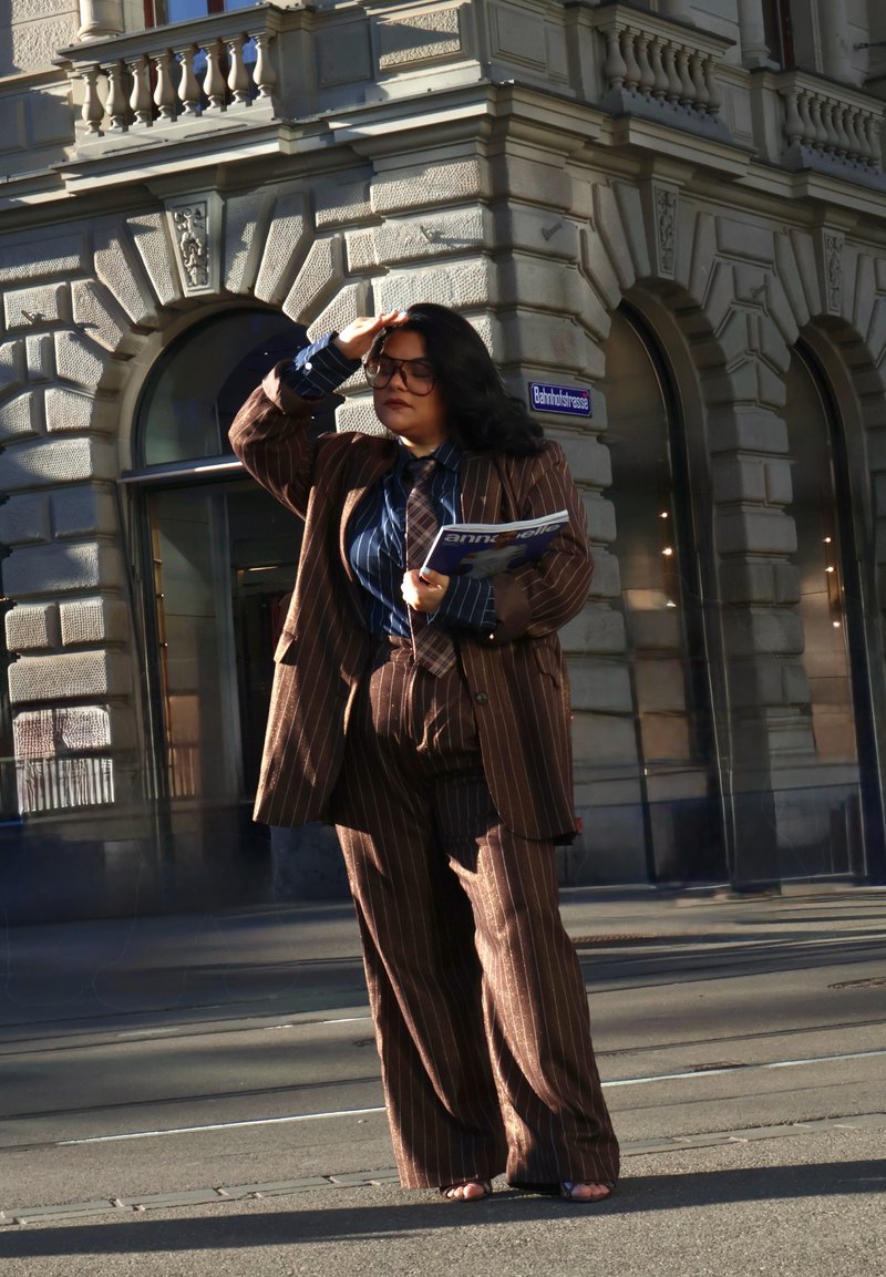 Femme en costume à fines rayures marron tenant un magazine, se protégeant les yeux du soleil, debout à un coin de rue avec une façade de bâtiment historique en arrière-plan.