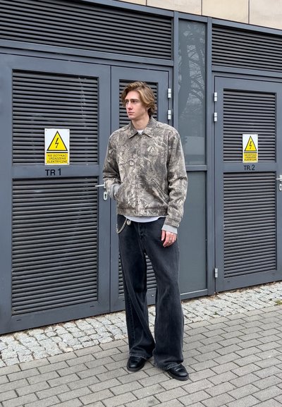 Chaqueta de camuflaje en beige y gris, combinada con pantalones de pierna ancha negros y zapatos negros. De pie al aire libre cerca de puertas con señales de advertencia.