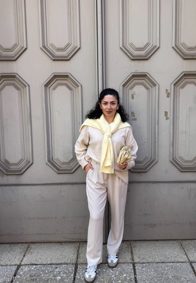 Suéter crema con una bufanda amarilla atada al cuello, pantalones beige y zapatillas blancas, de pie frente a una puerta gris texturizada.