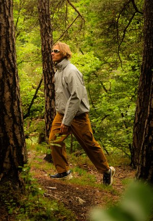 Giacca impermeabile grigio chiaro, pantaloni cargo arancioni con tasche e scarpe scure; persona che cammina su un sentiero forestale circondato da vegetazione.