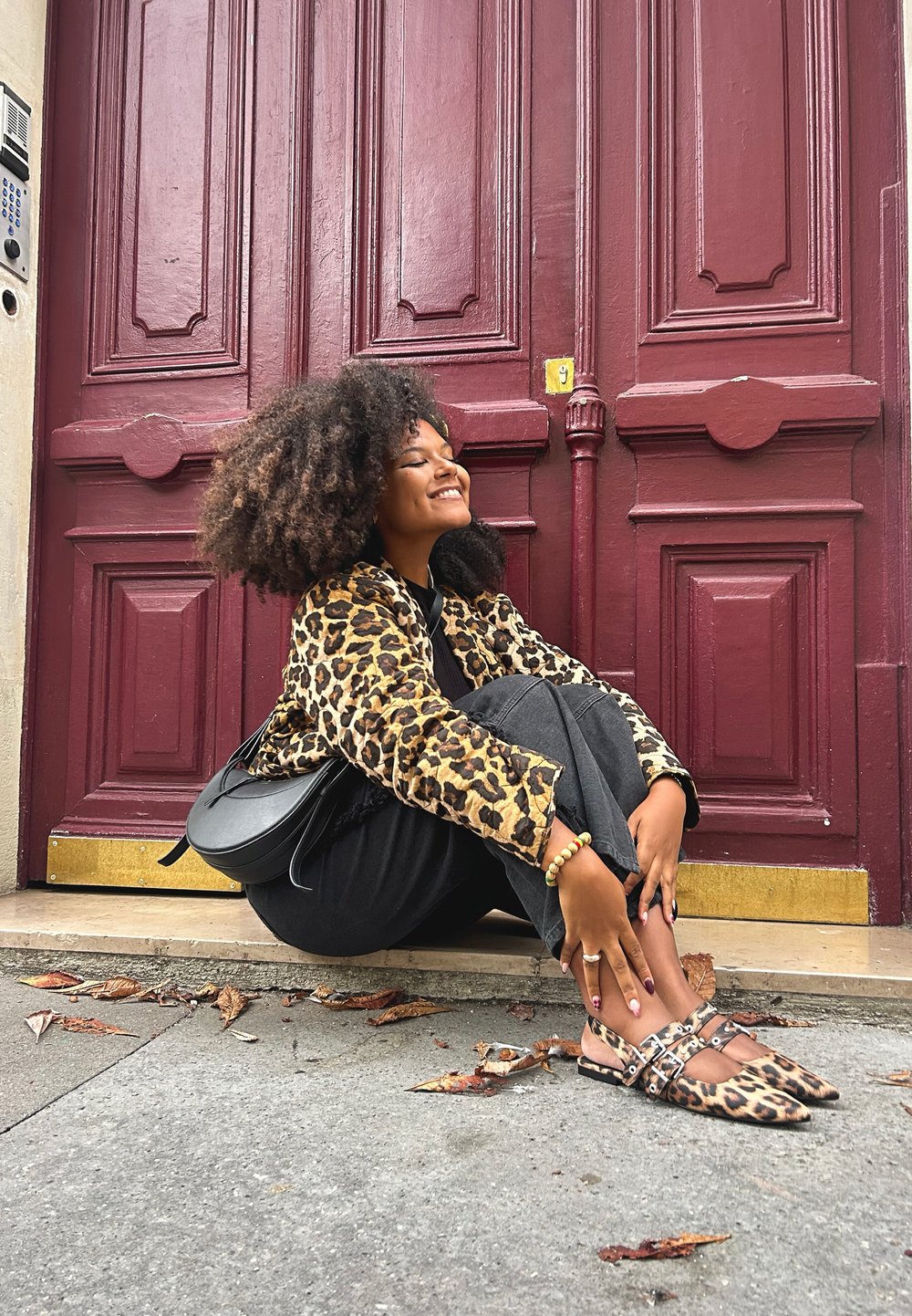 Leopardprint jas, zwarte kleding, schattige leopardprint schoenen met bandjes. Achtergrond heeft een bordeauxrode deur met gedetailleerde omlijstingen.