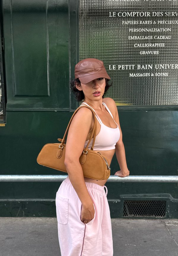 Donna con pantaloni rosa e top corto bianco, indossa un berretto marrone e una borsa a tracolla, in piedi e guarda di lato accanto a un muro verde con testo in francese.