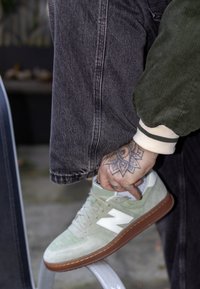 Baskets en daim vert avec logo blanc, semelle marron et perforations, associées à un jean gris et une veste verte texturée. Une main ajuste la chaussure.