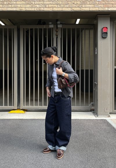 Una persona que lleva una camisa de cuadros en blanco y negro, una camiseta blanca, unos jeans anchos azules y zapatos de cuero marrón, sosteniendo un pequeño bolso marrón.