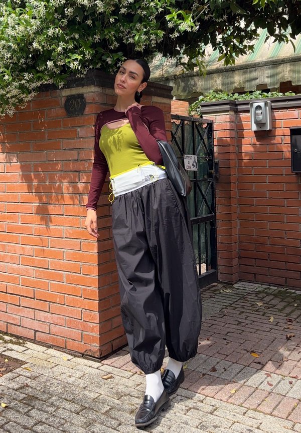 Frau lehnt an einer Ziegelmauer nahe einem Tor, trägt ein gelb-bordeauxfarbenes Oberteil, schwarze weite Hosen, weiße Socken und schwarze Loafer.
