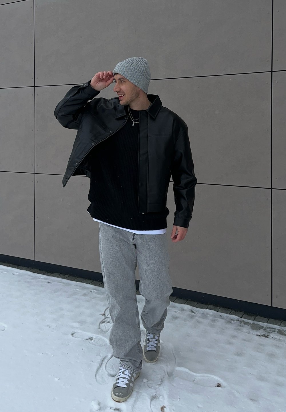Homme portant un bonnet gris, une veste noire, un pantalon gris et des baskets, marchant sur la neige à côté d'un mur de bâtiment, ajustant son chapeau avec un sourire.