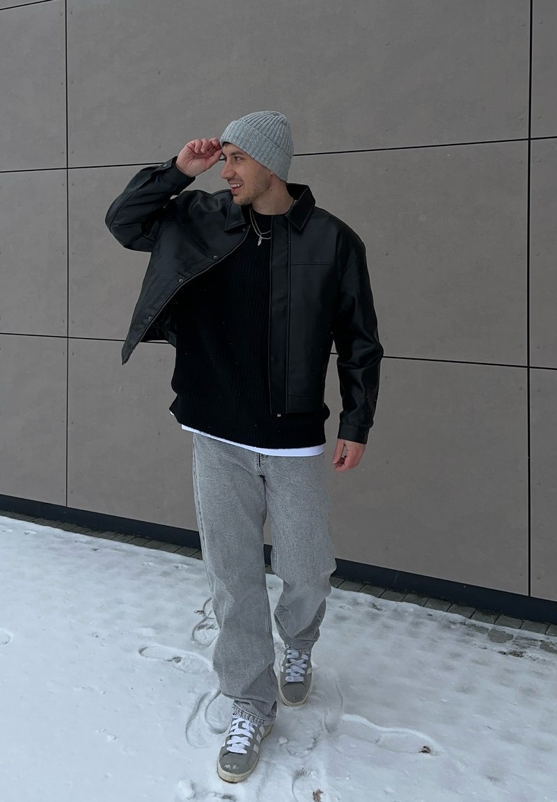 Homme portant un bonnet gris, une veste noire, un pantalon gris et des baskets, marchant sur la neige le long d'un mur de bâtiment, ajustant son chapeau avec un sourire.