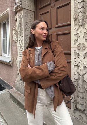 Joven mujer con el pelo largo y castaño, que lleva una chaqueta marrón, un jersey gris y pantalones blancos, está de pie con los brazos cruzados junto a la entrada ornamentada de un edificio.
