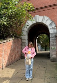 Ung kvinne i rosa jakke rekker inn i en beige håndveske nær en mursteinsbue dekket med graffiti og grønt.