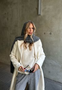 Manteau en laine blanche sur un pull beige, écharpe grise et pantalon de survêtement gris. Tenant des lunettes de soleil noires, avec des textures douces et des couleurs neutres.