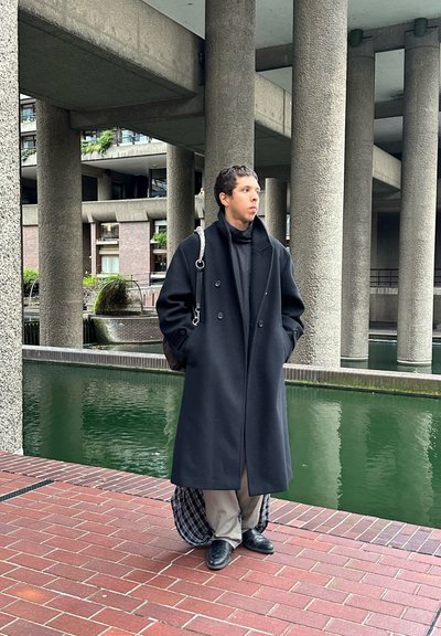 Abrigo negro de doble botonadura, tela texturizada, figura de pie, pantalones grises, zapatos de cuero, con un bolso de hombro, contra un camino de ladrillos y agua.