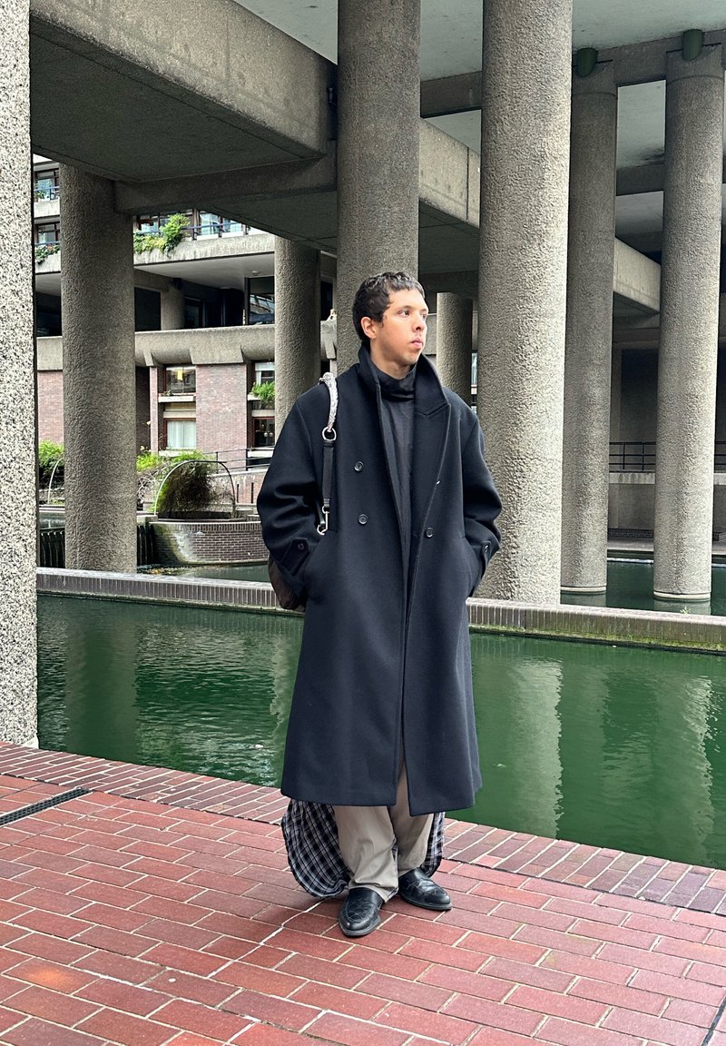 Manteau noir croisé, tissu texturé, silhouette debout, pantalon gris, chaussures en cuir, avec un sac à épaule, devant un chemin de briques et de l'eau.