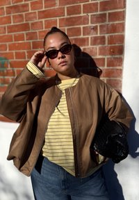 Brun bomberjakke, gulstripet skjorte, denim jeans. Tilbehør inkluderer svarte solbriller og en teksturert svart clutch. Mursteinsbakgrunn.