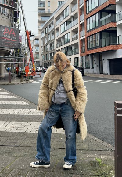 Abrigo de piel sintética en beige claro, corte relajado con mangas anchas, superpuesto a una sudadera gris, combinado con unos vaqueros sueltos azules y zapatillas Converse negras.