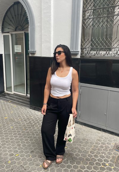 Top sin mangas blanco, pantalones negros ajustados, sandalias marrones. Sosteniendo un bolso de tote con estampado. El fondo presenta una fachada de edificio en blanco y negro.