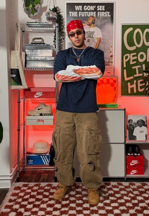 Hombre con bandana, gafas de sol, camisas superpuestas y pantalones cargo, de pie con los brazos cruzados en una habitación con cajas de zapatillas y carteles.