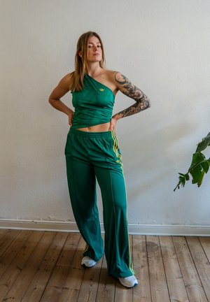 Mujer con tatuaje en el brazo, vestida con un top verde de un solo hombro y pantalones anchos a juego, de pie en el interior sobre un suelo de madera junto a una planta.