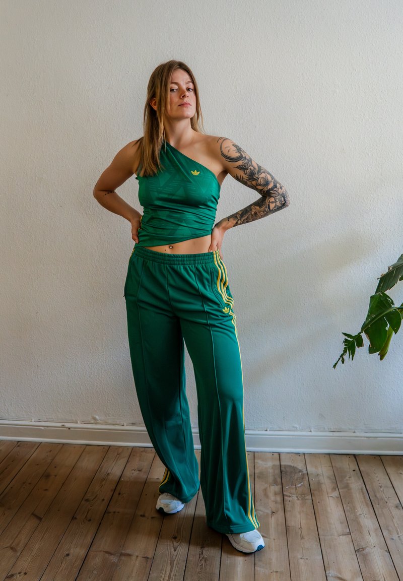 Femme avec un tatouage sur le bras portant un haut vert à une épaule et un pantalon large assorti, debout à l'intérieur sur un sol en bois à côté d'une plante.