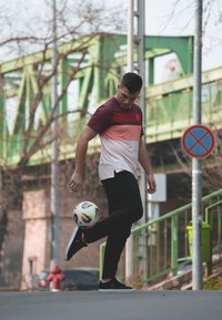 Trumpas rankovėmis, bordobordo ir rausvos gradientinės spalvos sportinė marškinėliai, juodos kelnės, atlieka kojų šokius su spalvingu futbolo kamuoliu gatvėje.